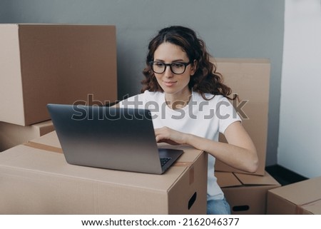 Girl in white t-shirt and glasses is texting on computer ordering delivery service. Happy hispanic woman packing boxes for moving to new place. Easy move concept. Royalty-Free Stock Photo #2162046377