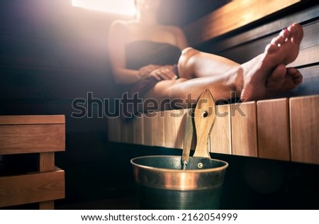 Sauna, steam room bath. Woman relaxing in spa. Wellness and warm temperature therapy in dark wood home in Finland. Water bucket and ladle. Towel on body, resting legs. Traditional Finnish lifestyle. Royalty-Free Stock Photo #2162054999
