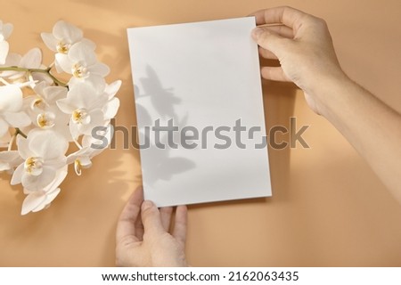 Hands are holding an empty white A5 form on a beige background. Layout top view Royalty-Free Stock Photo #2162063435