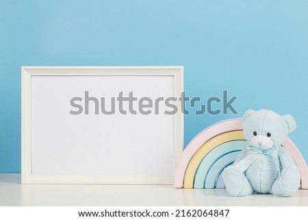 White empty wood picture frame with teddy bear and pastel wooden toy rainbow on white desk with light blue background. Front view