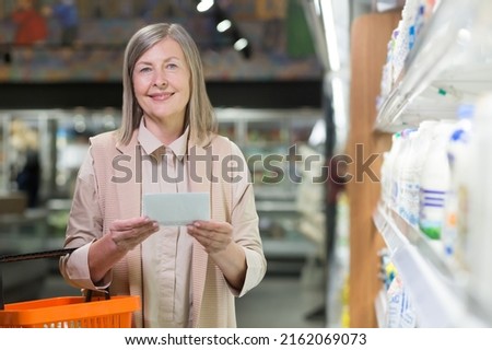 Portrait. Senior beautiful woman shopping in the store. Supermarket. Stands, holds a list of products written on paper in his hands, holds a plastic basket for products, chooses. looks at the camera