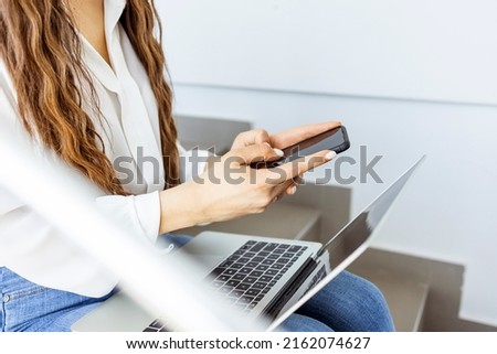 Portrait of young woman holding phone reading good news, while sitting over staircase at office. Young woman with laptop sitting on the stairs.  Person using personal computer outdoors