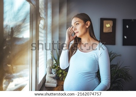 A young Caucasian pregnant woman with a headache is standing by the window in the living room. The concept of parenting expectations of pregnancy symptoms. A lady at home holding her baby's belly