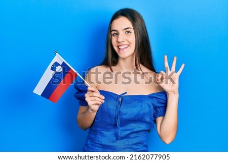 Young brunette teenager holding slovenia flag doing ok sign with fingers, smiling friendly gesturing excellent symbol 
