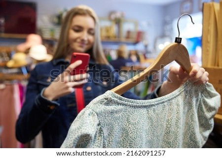 Young Woman Buying Used Sustainable Clothes From Second Hand Charity Shop Taking Picture Of Dress On Mobile Phone