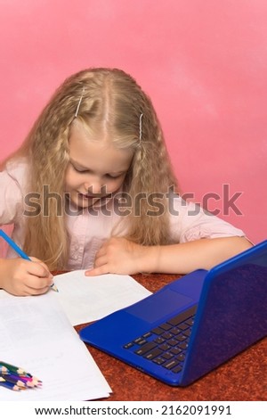 little schoolgirl with pencils writes at a table with a laptop. pink background