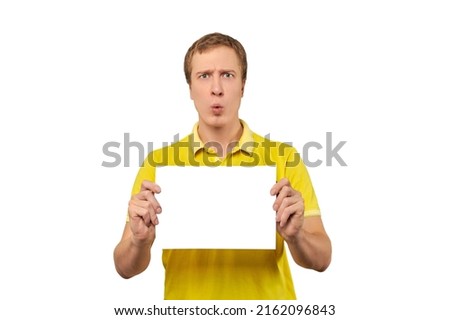 Surprised young man holding blank paper sheet, paper mockup isolated on white background. Funny amazed guy in yellow T-shirt with paper mock up, happy excited emotions and pleasantly shocked face