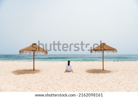 Back view of anonymous lady sitting on sandy shore near straw umbrellas. She is enjoying summer vacation while contemplating waving sea under blue sky