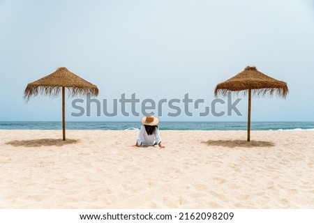 Back view of anonymous lady sitting on sandy shore near straw umbrellas. She is enjoying summer vacation while contemplating waving sea under blue sky