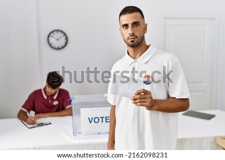 Young hispanic man voting putting envelop in ballot box thinking attitude and sober expression looking self confident 
