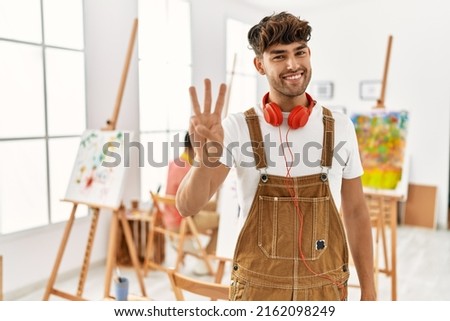 Young hispanic man at art studio showing and pointing up with fingers number three while smiling confident and happy. 