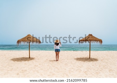 Back view of anonymous lady standing on sandy shore near straw umbrellas. She is enjoying summer vacation while contemplating waving sea under blue sky