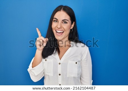 Young hispanic woman standing over blue background pointing finger up with successful idea. exited and happy. number one.  Royalty-Free Stock Photo #2162104487