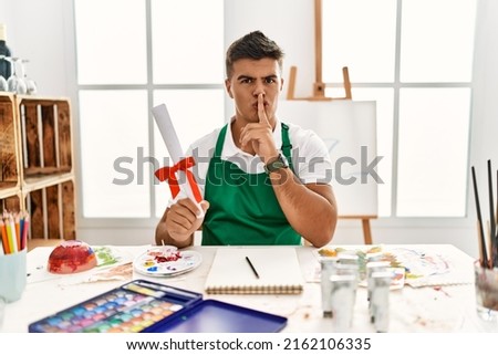 Young hispanic man at art studio holding degree asking to be quiet with finger on lips. silence and secret concept. 