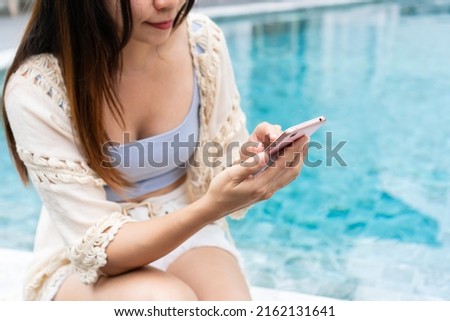 Young Asian Woman using mobile phone and sitting besides swimming pool. Beautiful traveler relaxes and uses smart phone besides water pool of hotel. Summer vacation, technology and lifestyle concept.
