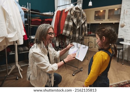 Pleasant mature European woman, cheerful caring and loving grandma admiring the drawing - clothes sketch- of her granddaughter adorable baby girl while working in a fashion design workshop