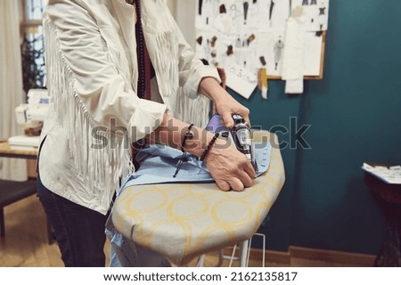 Detail of tailor's hands using steamer iron ironing clothes on ironing board in fashion design and tailoring atelier on the background of blurred sketches pinned on wall and sewing machine on a desk