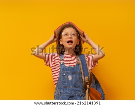 Cute happy kid standing on bright yellow background. Child with backpack and book, little girl is ready to back to school. 