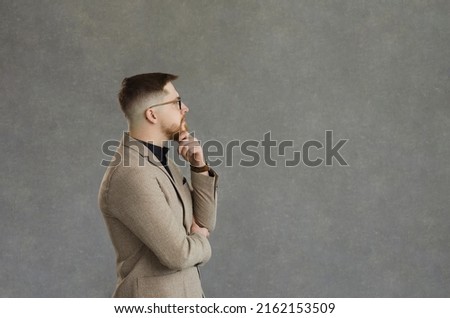 Profile portrait of serious man in jacket standing and looking at text copyspace on grey background. Side view young business professional thinking hand on chin about interesting quote or project idea