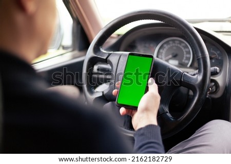 A man is sitting in a car and holding a smartphone with a green screen on the steering wheel. The concept of an application for taxi drivers
