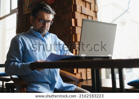 Male freelancer is working in a cafe on a new business project. Sits at a large window at the table. Looks at a laptop screen with a cup of coffee.