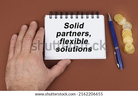 Business concept. On a brown surface are coins, a pen and a notepad with the inscription - Solid partners flexible solutions. The notebook is holding a hand.