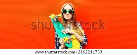 Portrait of modern young blonde woman with green skateboard wearing sunglasses on colorful red background