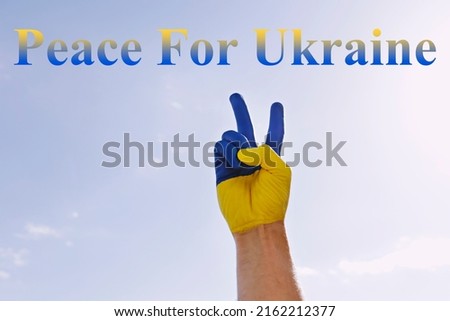 Peace for Ukraine slogan written over blue sky background. Man's hand painted in colors of Ukrainian national flag showing a peace gesture as sign of support and solidarity with Ukraine. Copy space.