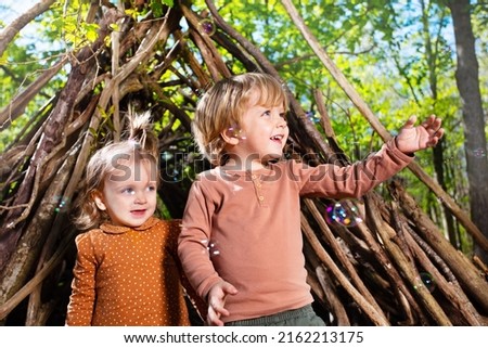 Happy little kids brother and sister girl play with soap bubbles in hut of branches in the forest