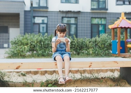 little cute girl 4 years old sits on a bench on the playground and looks at the phone, plays or watches a cartoon, the concept of children's dependence on gadgets