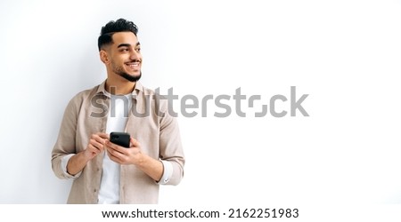 Panoramic photo of a joyful handsome indian or arabian guy, holding smartphone in hand, chatting online, browsing internet, looking happily to the side, standing on isolated white background, smiling Royalty-Free Stock Photo #2162251983