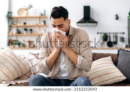 Unhappy sad indian or arabian guy suffering from fever and flu, sitting on sofa in living room, blowing nose and sneezing in napkin, need treatment of illness, and doctors consultation Royalty-Free Stock Photo #2162252043