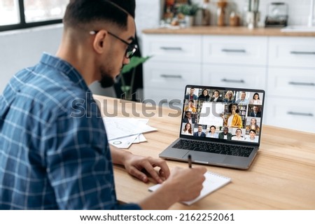 Distance learning, online course. View over the shoulder of a guy taking notes in a notebook, at a laptop screen with a teacher and a group of multiracial students. E-learning, webinar, online lecture Royalty-Free Stock Photo #2162252061