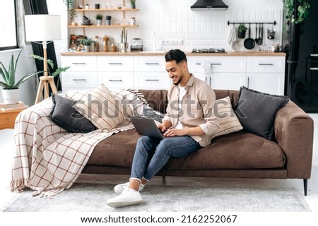 Positive handsome smart indian or arabian guy, freelancer or student, work or study online from home, using laptop, while sit on comfortable sofa in cozy living room, browsing internet, news, smiling Royalty-Free Stock Photo #2162252067