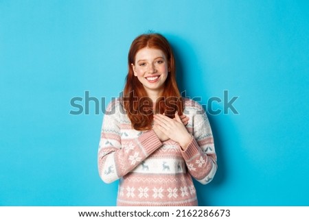 Image of beautiful redhead female model holding hands on heart and smiling, saying thank you, being grateful, standing over blue background