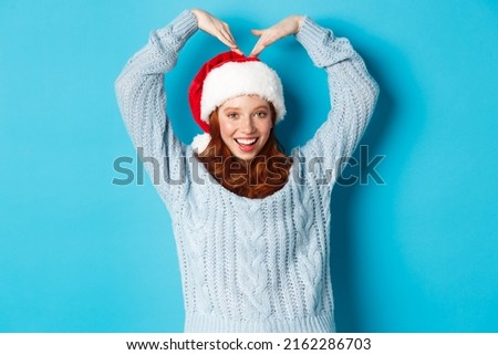 Winter holidays and Christmas Eve concept. Cute redhead teen girl in santa hat and sweater, making heart sign and smiling, wishing merry xmas, standing over blue background