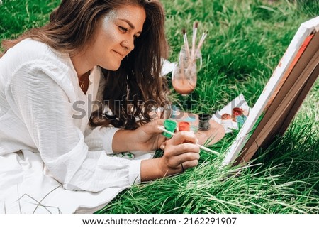 Artist painting on the easel outdoors in the garden. Open air outdoor art workshop. Draw on the canvas with brush and palette sitting on the grass during a picnic in a park