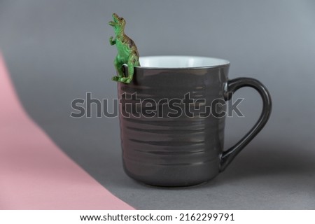 Miniature of a dinosaur sitting on a dark gray mug.  Small green figurine of a predatory dinosaur. Ceramic mug with handle for hot drinks. Gray-pink background. Selective focus.