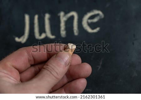 Adult male holding a piece of yellow chalk in his hand. Handwritten word JUNE on black chalkboard. Selective focus.