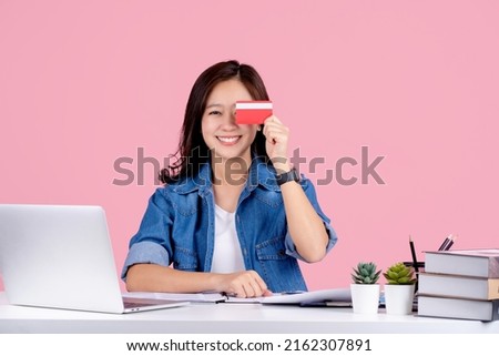 Young asian business woman in casual shirt sit work at white office desk with pc laptop.  She cover eye with credit bank card isolated on pastel pink background.