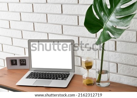 Workplace with laptop, hourglass, clock and palm leaf in vase near white brick wall