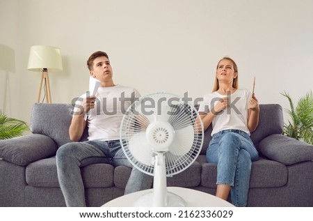 Young couple at home suffering from hot sultry sweltering summertime weather. Sweaty exhausted people waving sheets of paper sitting on sofa with electric fan in overheated house. Summer heat concept Royalty-Free Stock Photo #2162336029