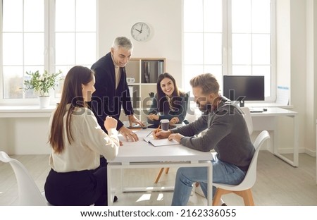 Group of positive people dressed in casual clothes discuss plan for cooperation on common project in modern coworking. People write their ideas in notebook sitting at table in bright office. Royalty-Free Stock Photo #2162336055