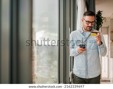 Young businessman analyzing credit card while using smart phone. Professional is making online payment. He is standing by window at office.