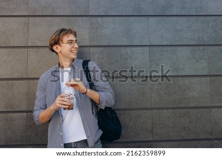 Outdoor portrait of handsome guy standing in park with coffee, and looking aside. Man wear stylish clothes and glasses
