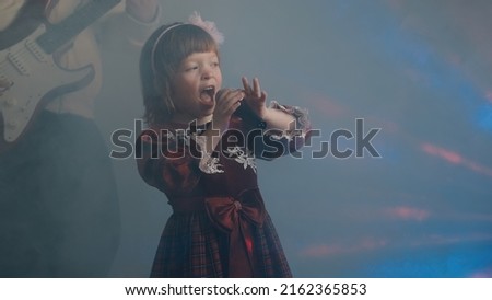 Little girl on stage in vintage dress, she sings into microphone and dances, her father plays an electric guitar. Color music is shining and smoke is billowing. Family time, creativity and hobbies
