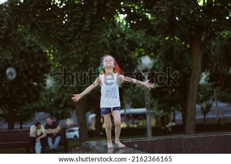 pretty little girl with colorful dyed hair holding bunch of baloons running along fountain parapet. Image with selective focus