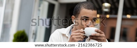 african american man in wireless earphones drinking coffee on summer terrace, banner