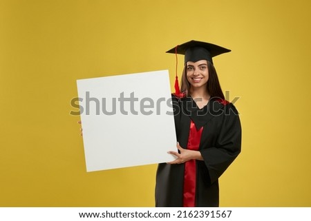 Front view of girl in mortarboard standing, smiling, looking at camera. Pretty young female graduating from college, holding, showing. Isolated on yellow studio mockup background.