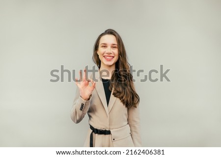 Positive female office worker in suit stands on beige background with smile on face and shows OK gesture with fingers.